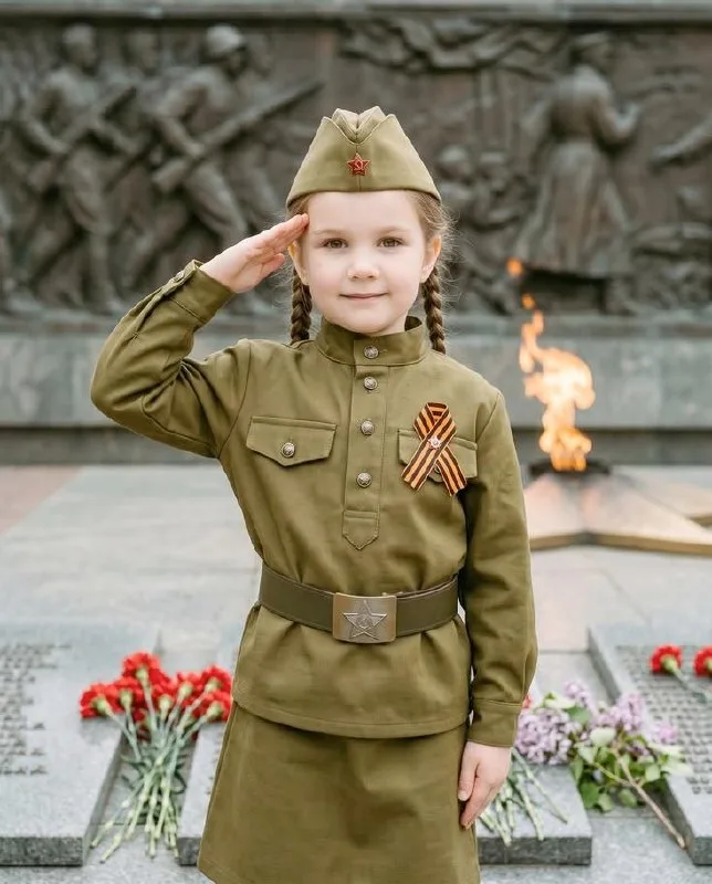 Девочка в детской гимнастёрке и пилотке с косичками отдаёт честь у горельефа памятника и Вечного огня.