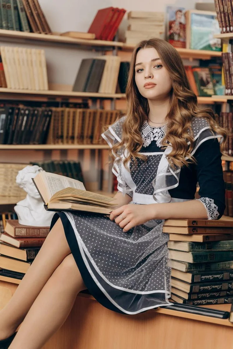 Классический портрет выпускницы в школьной форме: коричневое платье с белым фартуком, мягкие локоны, сидит на стопке книг в библиотеке, в руках раскрытый том, рядом бюст Пушкина.